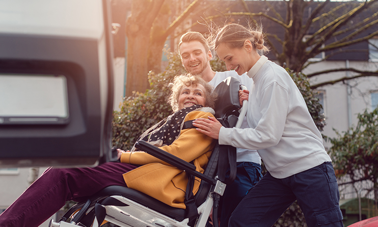 woman in a wheelchair being helped into transport by two carers.