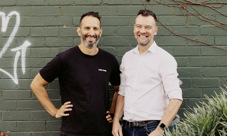 Two men stand smiling against a brick wall