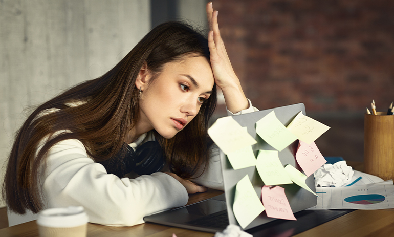 woman sat at desk with laptop covered in postits looking fatigued, burnout
