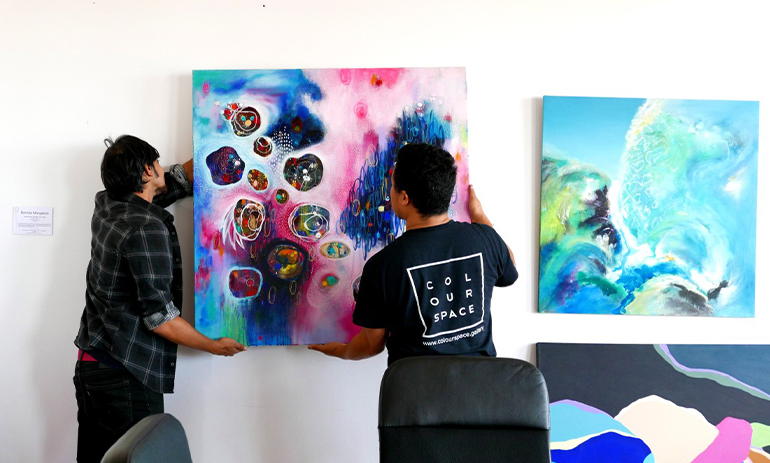 Two people hanging a colourful painting on a white wall