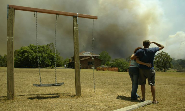 couple standing next to a swing looking at smoke from a bushfire behind a hosue