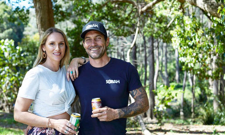 a man and a woman stand under a tree holding a can of non alcoholic beer