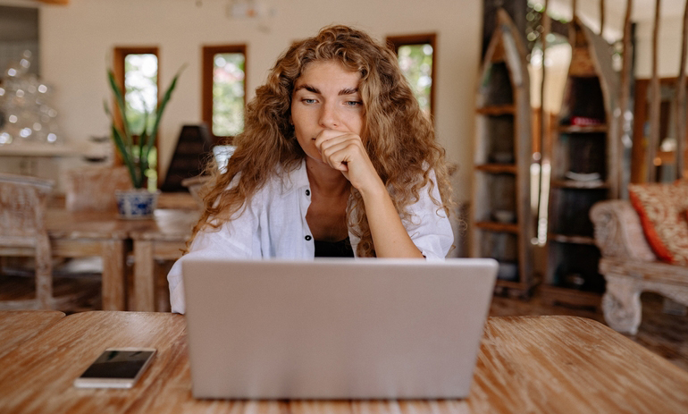 woman with long curly hair sitting at a table looking at a laptop