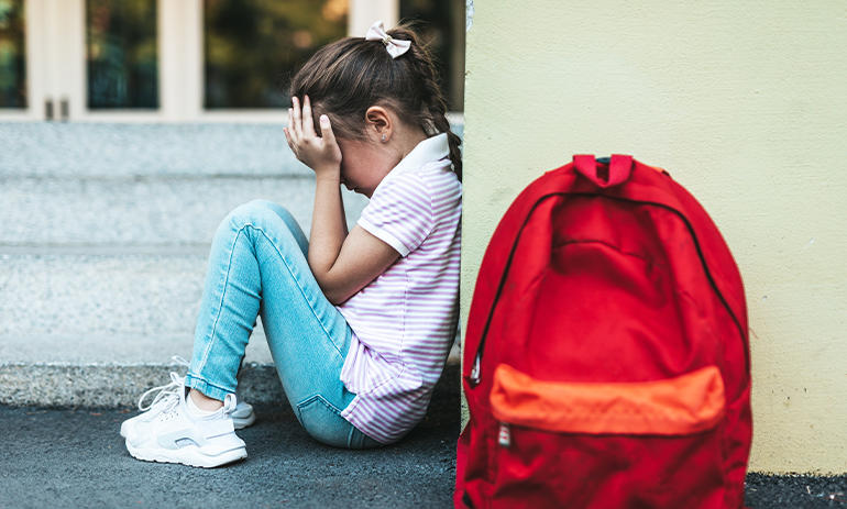 young girl sitting on the floor crying with a red backpack in the foreground