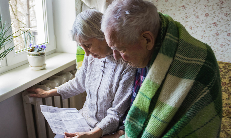 older couple wearing a blanket witting in front of the radiator looking at a bill
