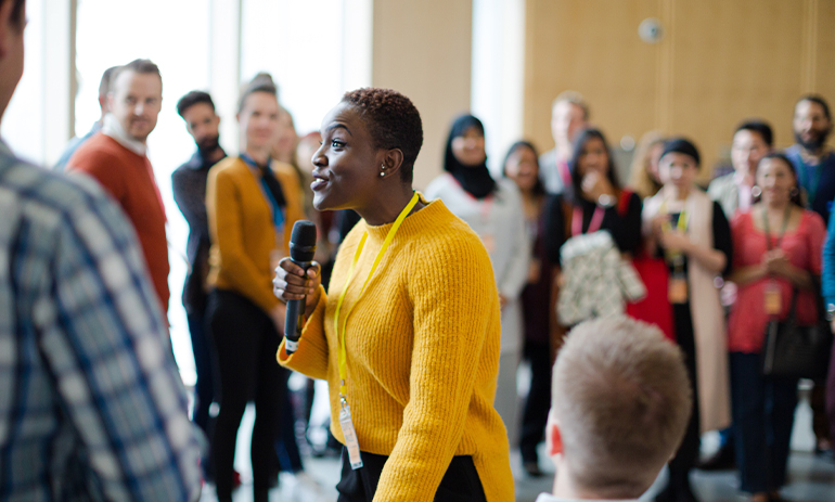 black woman in yellow jumper speaking to a large group of people