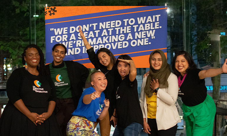 group of Bla(c)k women and women of colour in front of a sign that says "we dont need to wait for a seat at the table, we're making a new and improved one".