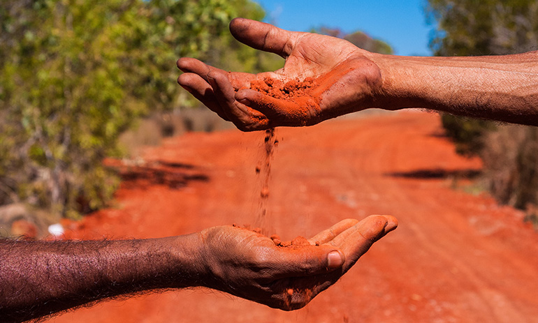 A hand pours sand into another hand against an outback backdrop.
