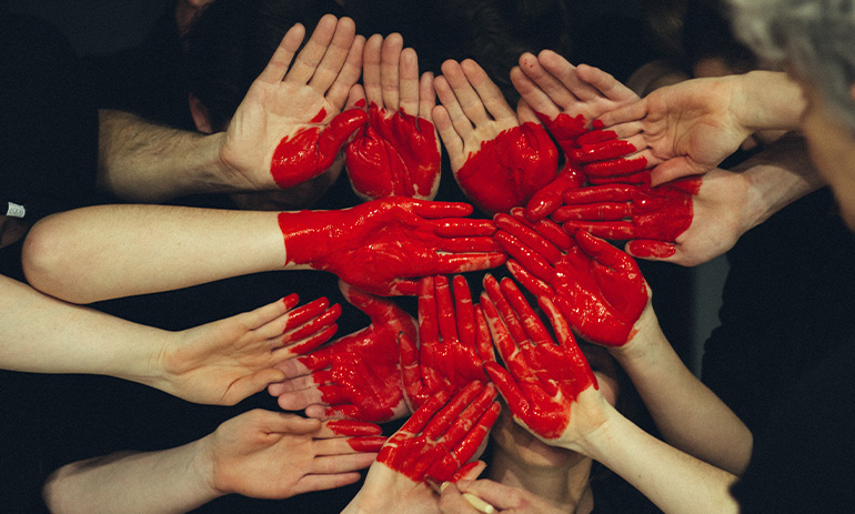 A group of people with their hands painted red put them together in the shape of a heart.
