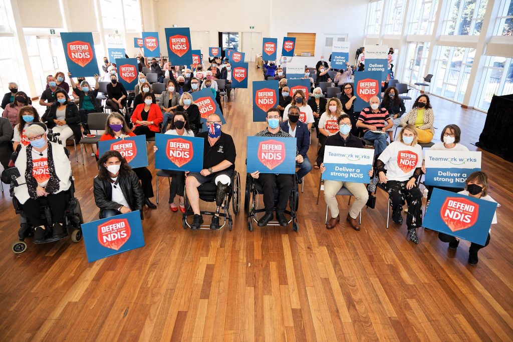 A group of protestors holding signs gather to Defend the NDIS