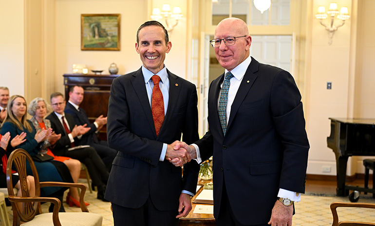 Andrew Leigh, wearing a black suit and red tie, shakes hands with Governor General David Hurley, who is an older man in a black suit and dark tie, while being sworn in as assistant minister.