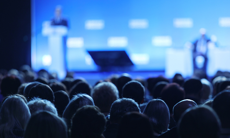 audience at a conference lookind at a speaker on the stage, the stage is blurred and tinged blue