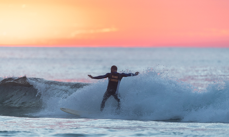 An image of someone surfing with a pink sunset in the background. Text on their wetsuit reads: “Blind Surfer”.