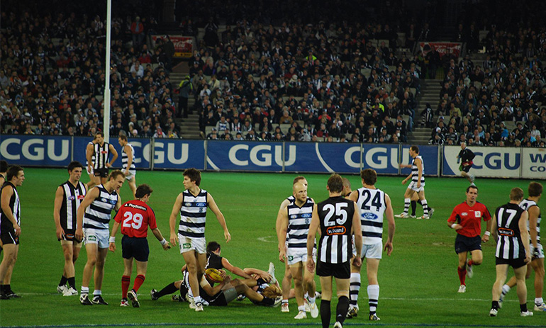 A group of AFL players on field. Some are wearing the black and white stripes of Collingwood, others are wearing the blue and white stripes of Geelong. There is a crowd watching them in the stands in the background. The signs around the ground have the logo of insurance company CGU, which is a blue background with big white letters saying CGU.