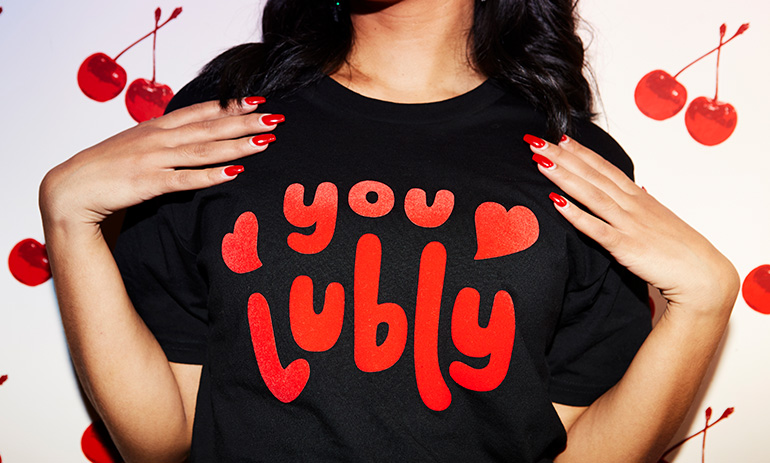 A lady wears a black tshirt that features red writing that says 'you lubly'. She has red nail polish on and dark hair. We can only see her torso.
