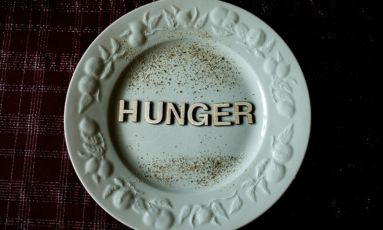 A white plate with a decorative border, sits on a dark background. On the plate are some grains and the word Hunger.