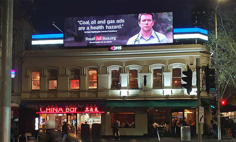 A city street, featuring two restaurants with a billboard above them. The billboard has a picture of a male doctor on it, and the words 'coal, oil and gas ads are a health hazard'.