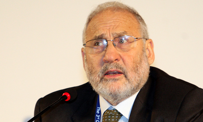 An older man is looking sideways, away from the camera. He has glasses and grey balding hair and a grey beard. He is wearing a black suit and white shirt with a dark coloured tie. He looks concerned.