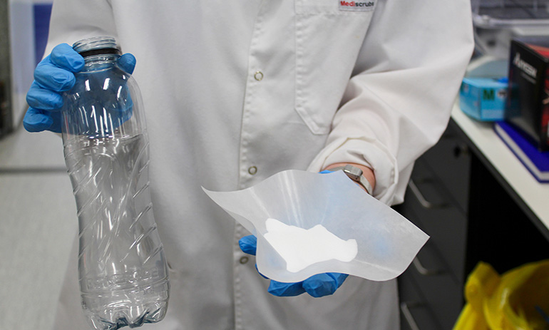 A person in a white lab coat, wearing blue gloves, stands front on to the camera holding a clear plastic bottle and a bowl of a white, powdery substance