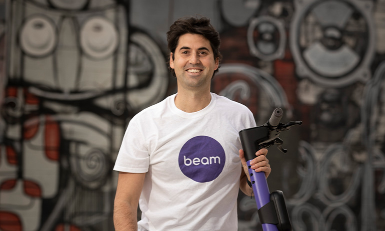 Tom, the general manager at Beam, is a white man with dark hair. He is smiling and wearing a white tshirt with a purple circle logo on it that says Beam. He is holding the handlebars of a purple scooter.