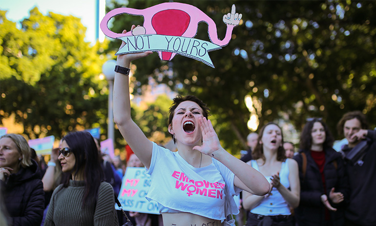 A young woman shouts at the camera. She has spiky brown hair and is wearing a white midriff top. She is holding a poster in the shape of a uterus that says 'not yours'. Her shirt says 'empower women'.