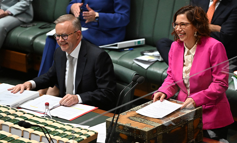 Anthony Albanese, the Prime Minister of Australia, sits on the left of the image at a table in the House of Representatives, with lots of papers in front of him. Amanda Rishworth, the social services minister, stands next to him and is smiling brightly. She is wearing a pink blazer and has dark curly hair.
