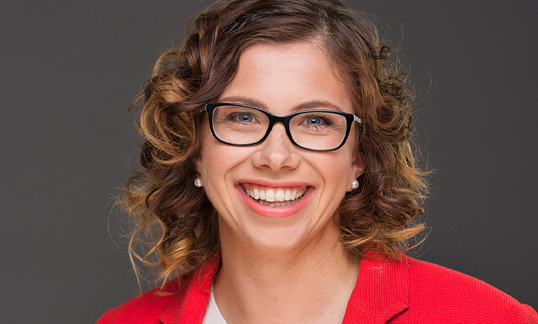 Amanda Rishworth smiles at the camera. She is a white lady with curly brown hair and black-rimmed glasses. She wears a red blazer.