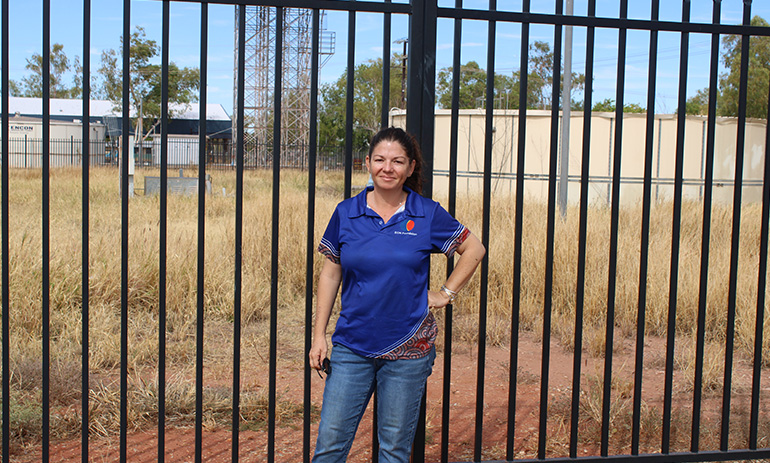 A lady stands in front of a black fence which is around a grassy space. She is wearing a blue polo shirt and jeans and has dark hair.