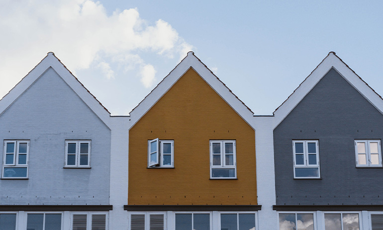A row of three houses with pointed roofs, one white, one mustard yellow and one grey, under a sunny sky.