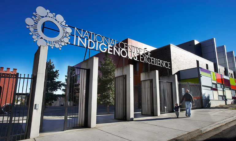 The National Centre of Indigenous Excellence, a cluster of square buildings on a street, with five grey pillars at its entrance