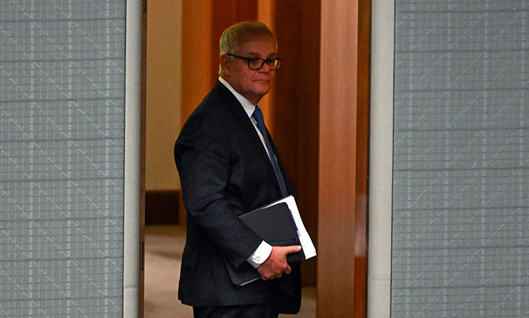 Former Prime Minister Scott Morrison stands framed in a doorway, half in shadow. He is carrying a compendium of papers and wearing a dark suit and glasses. The picture looks almost sinister.