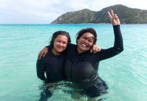 Two women with dark skin and hair are floating in a blue bay. They are in wetsuits and are smiling and having fun.