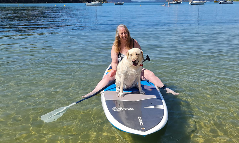 A lady sits on a paddleboard in the water with her guide dog. She is smiling and the dog is wet.