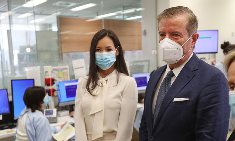 A well-dressed man and woman stand in front of researchers on computers. The lady has dark hair and is wearing a cream coloured outfit. The man has grey hair and is in a dark suit. They are both wearing N95 masks.