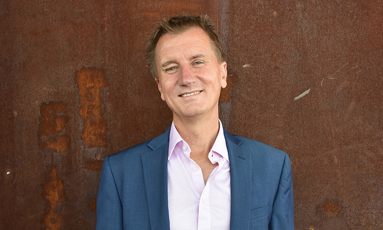Professor Nick Klomp smiles at the camera. He is wearing a blue suit and standing against a rust coloured background.