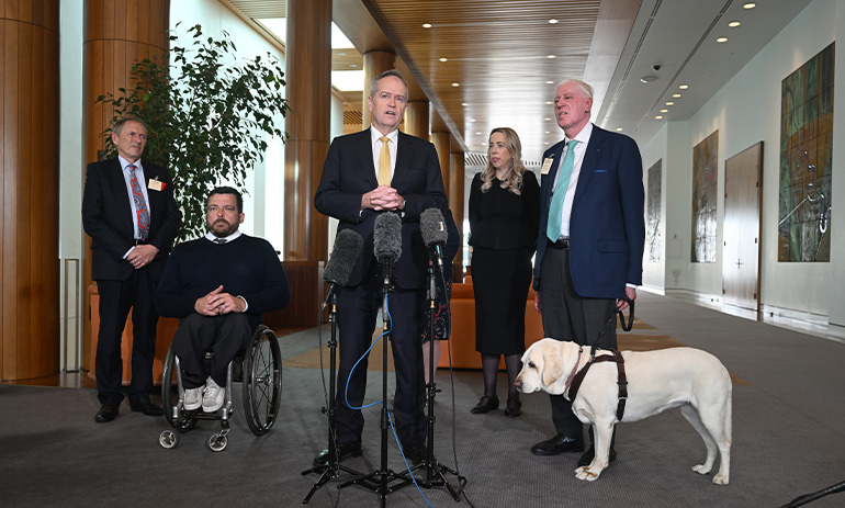 Five people stand in a Parliament House hallway. In the centre is NDIS Minister Bill Shorten. To his right is Kurt Fearnley, in a wheelchair. On his left are Rebecca Falkingham and Graeme Innes with a white guide dog.