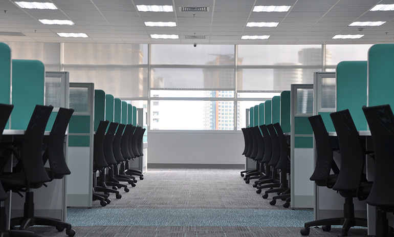 The inside of an office, featuring rows of black desk chairs in blue cubicles. You can see buildings through the window.