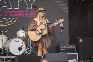 Madi Colville-Walker, in a dress and hat, plays an acoustic guitar on stage.