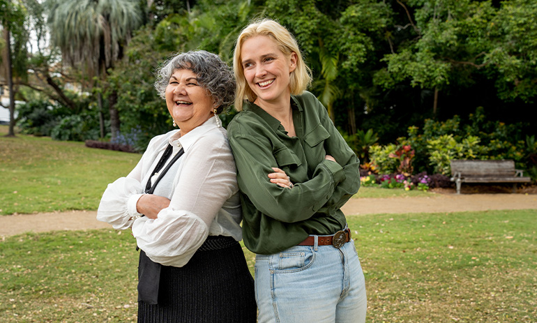 Two women stand back to back, their arms folded. They are laughing. On the left is a First Nations woman and on the right is a white woman with blonde hair.
