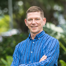 A headshot of Steve Williams, who has brown hair, is wearing a blue shirt and standing in front of a lush green garden.