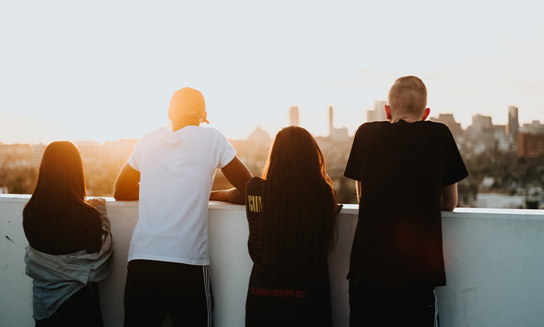 four young people stand on a balcony, their backs to the camera, watching the sunset over a city.