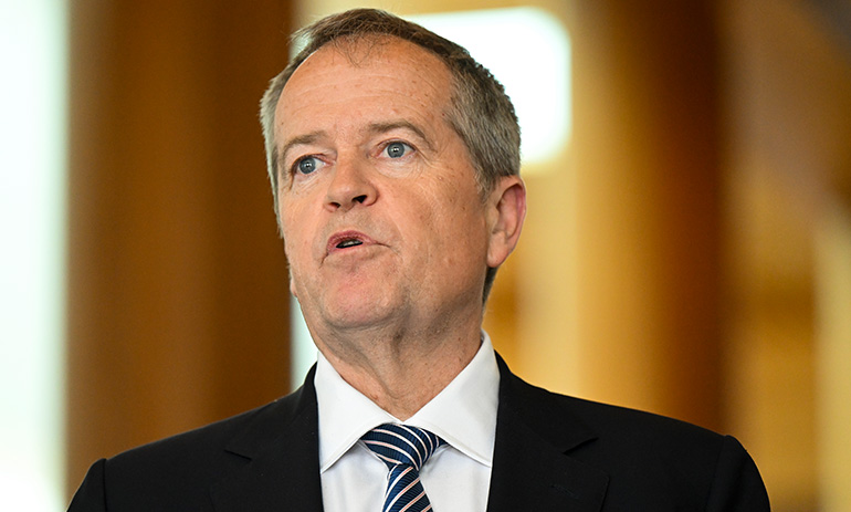 NDIS Minister Bill Shorten, making a speech. He is wearing a white shirt, blue tie and black jacket.