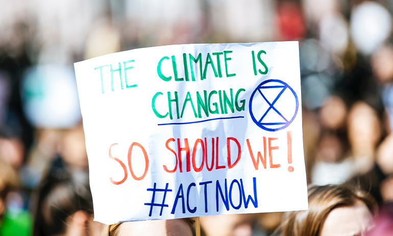 A poster being held at a rally, with colourful text reading: 'The climate is changing, so should we! #ActNow'