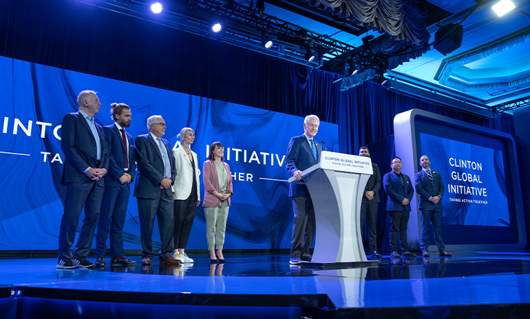 The image shows a group of people on a stage at a conference, including former US President Bill Clinton who is standing behind a lectern.