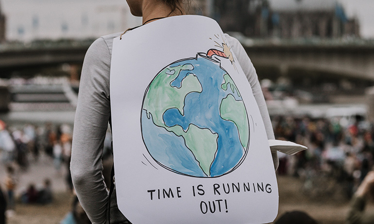 A girl stands with her back to the camera. Over her backpack is a sign with a hand drawn picture of the earth that says 'time is running out'
