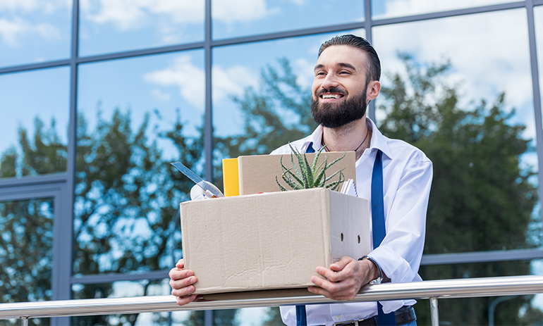 Man with beard standing outside office holding a box of items