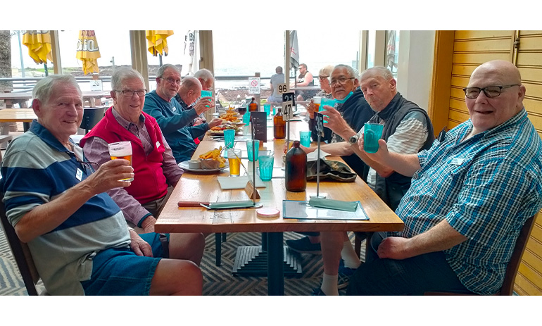 A group of men sit at a table at a pub. They are all smiling and raising a glass to the camera.