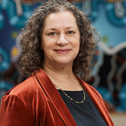 Kelly Bruce smiles at the camera. She has brown curly hair and wears an orange top.