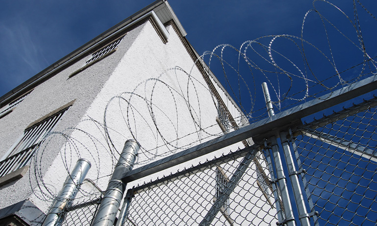 A prison fence topped with spiral barbed wire, with a prison building behind. We are looking up at the fence and building.