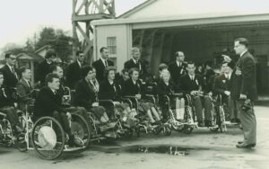 A black and white photo of a group of people in wheelchairs, being spoken to by a man in a suit. They are all well dressed.